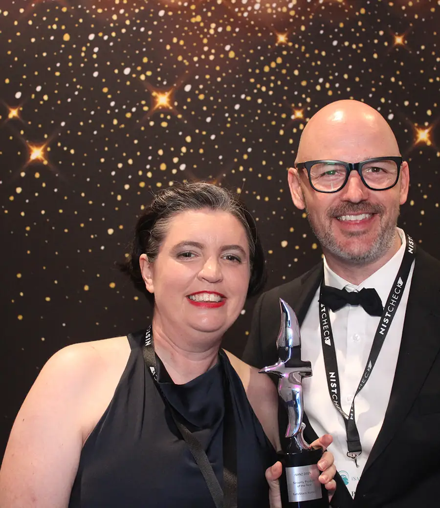 Two guests in evening wear holding an award trophy in front of a gold sparkle backdrop