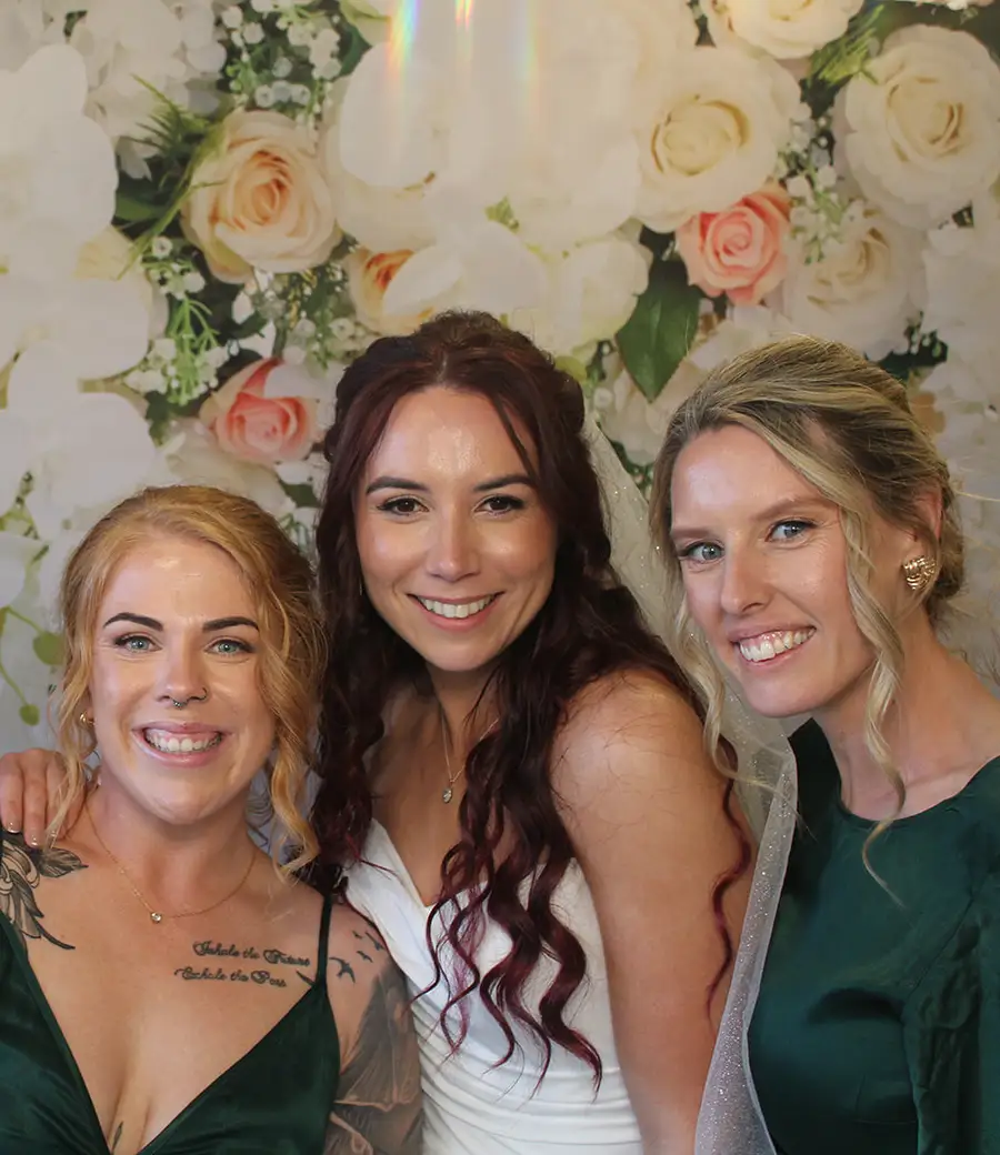 A bride and two bridesmaids in green dresses smiling in front of a white flower wall
